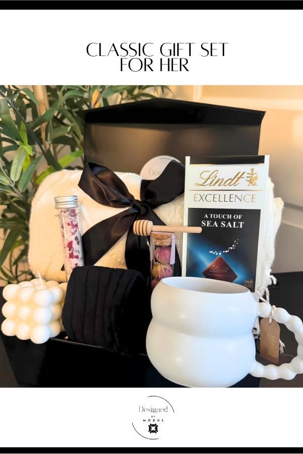 Elegant holiday gift box for her featuring cozy blanket, mug, candle, socks, tea, bath salts, and chocolate presented in a black box with satin bow.