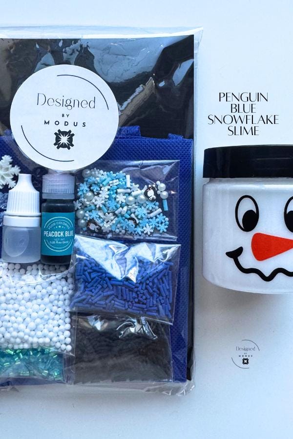 Packaged Penguin Blue Snowflake Slime Kit with Designed by Modus label, navy apron, snowman jar, and blue winter-themed mix-ins labeled “Penguin Blue Snowflake Slime.”