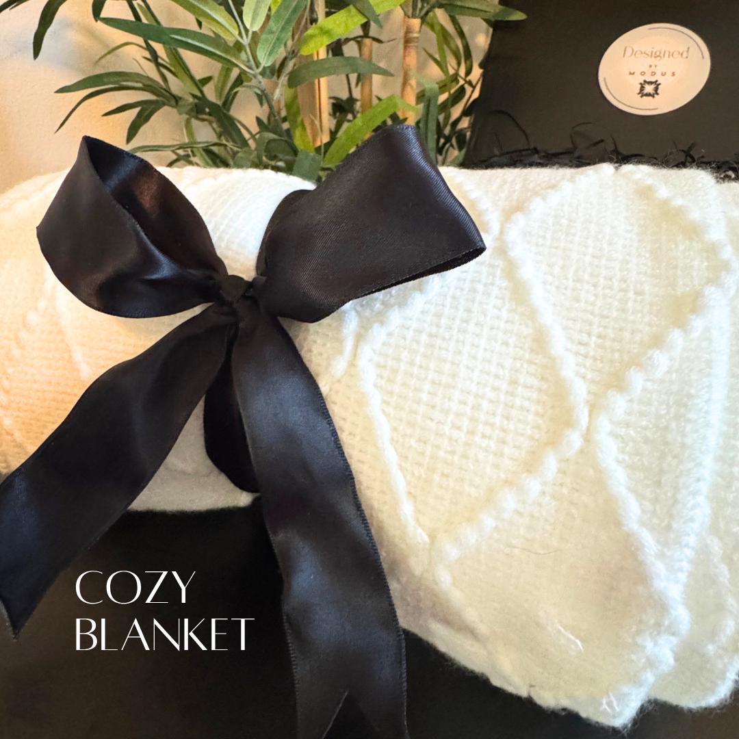 Soft white knit throw blanket tied with black satin ribbon, part of luxury gift set for women.