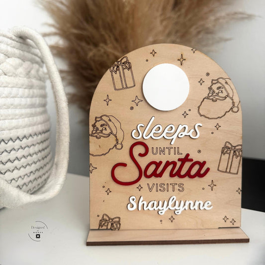 Personalized wooden “Sleeps Until Santa” countdown sign with engraved Santa faces and gift illustrations on black and white checkered background.