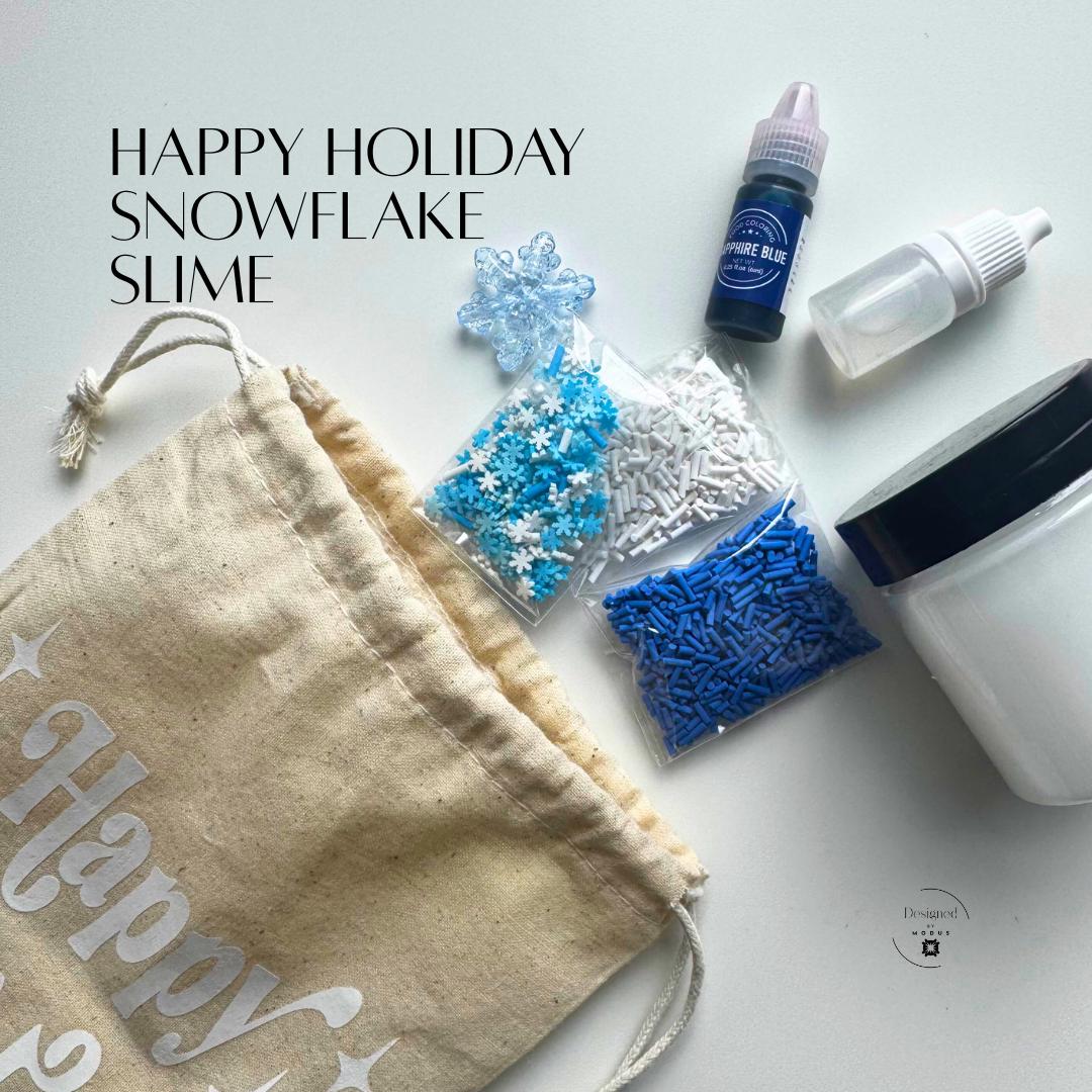 Flat lay of Happy Holiday Snowflake Slime Kit with blue dye, snowflake sprinkles, and clear activator bottle beside beige linen bag with white lettering.