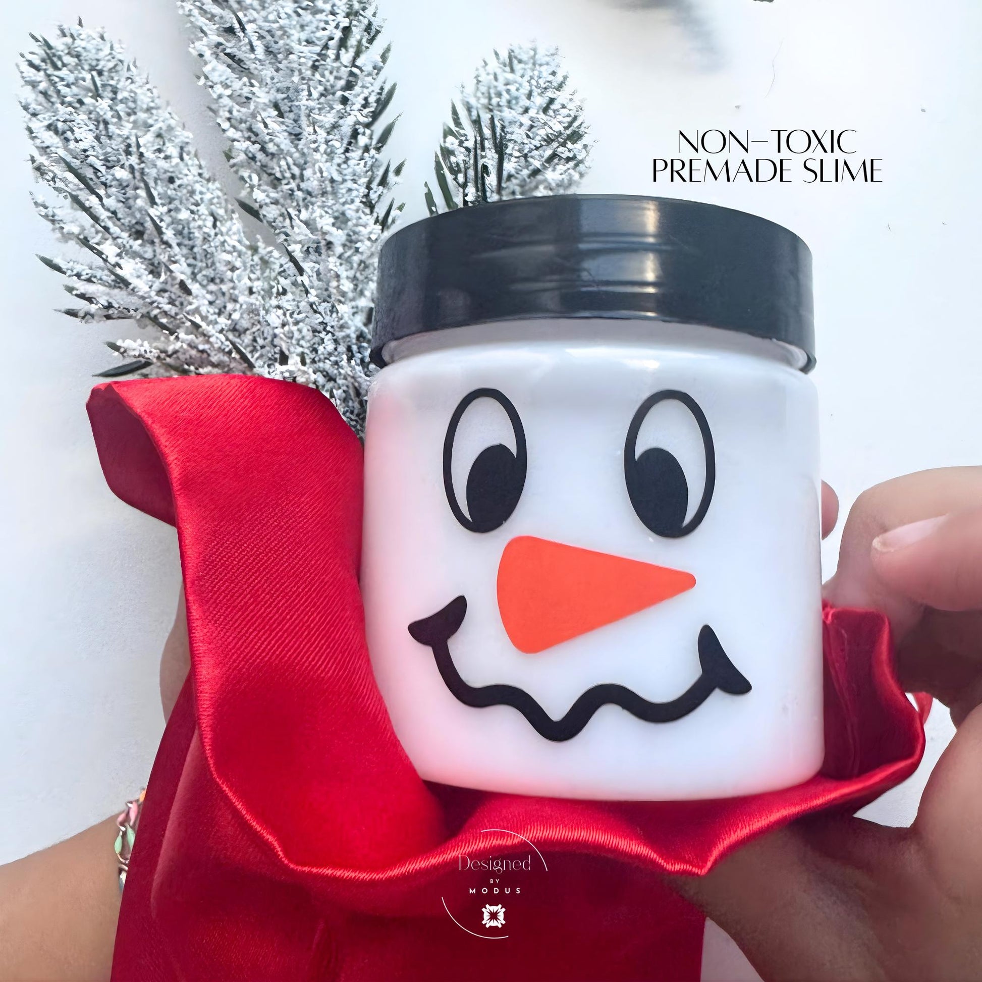 Non-toxic premade white slime in snowman jar with black lid, wrapped in red satin ribbon with frosted holiday greenery for Christmas gifting.