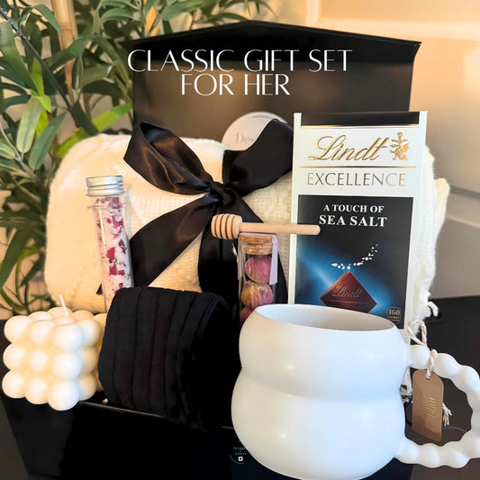 Elegant holiday gift box for her featuring cozy blanket, mug, candle, socks, tea, bath salts, and chocolate presented in a black box with satin bow.