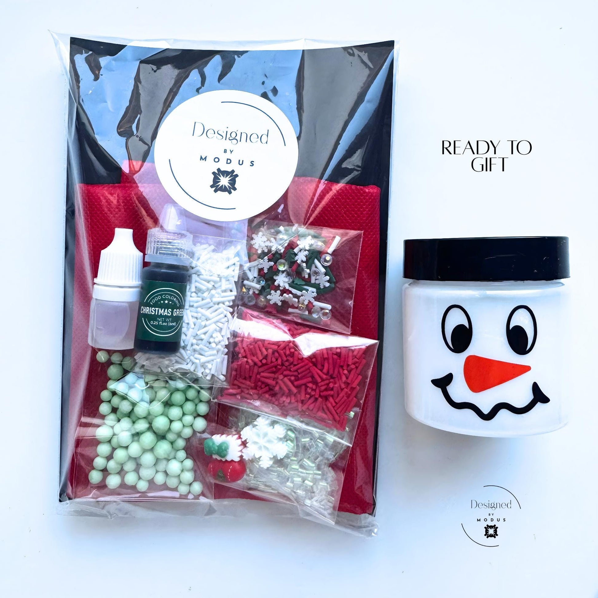 Packaged in clear plastic -Christmas Tree Slime Kit with red apron, pre-made white slime in snowman jar, green dye, activator bottle, and assorted red, green, and white holiday mix-ins including sprinkles, foam beads, and charms displayed flat lay style.
