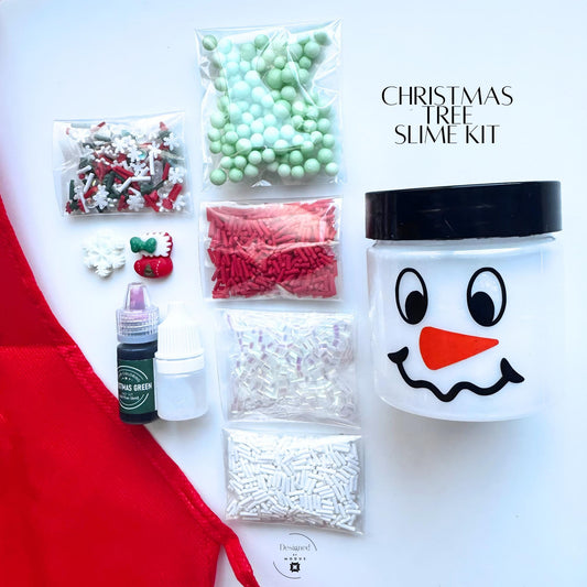 Christmas Tree Slime Kit with red apron, pre-made white slime in snowman jar, green dye, activator bottle, and assorted red, green, and white holiday mix-ins including sprinkles, foam beads, and charms displayed flat lay style.