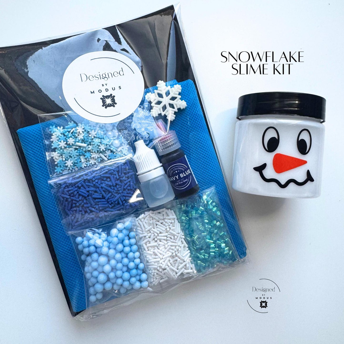 Packaged Snowflake Slime Kit with Designed by Modus label, blue apron, snowman jar, and winter-themed mix-ins labeled “Snowflake Slime Kit.”
