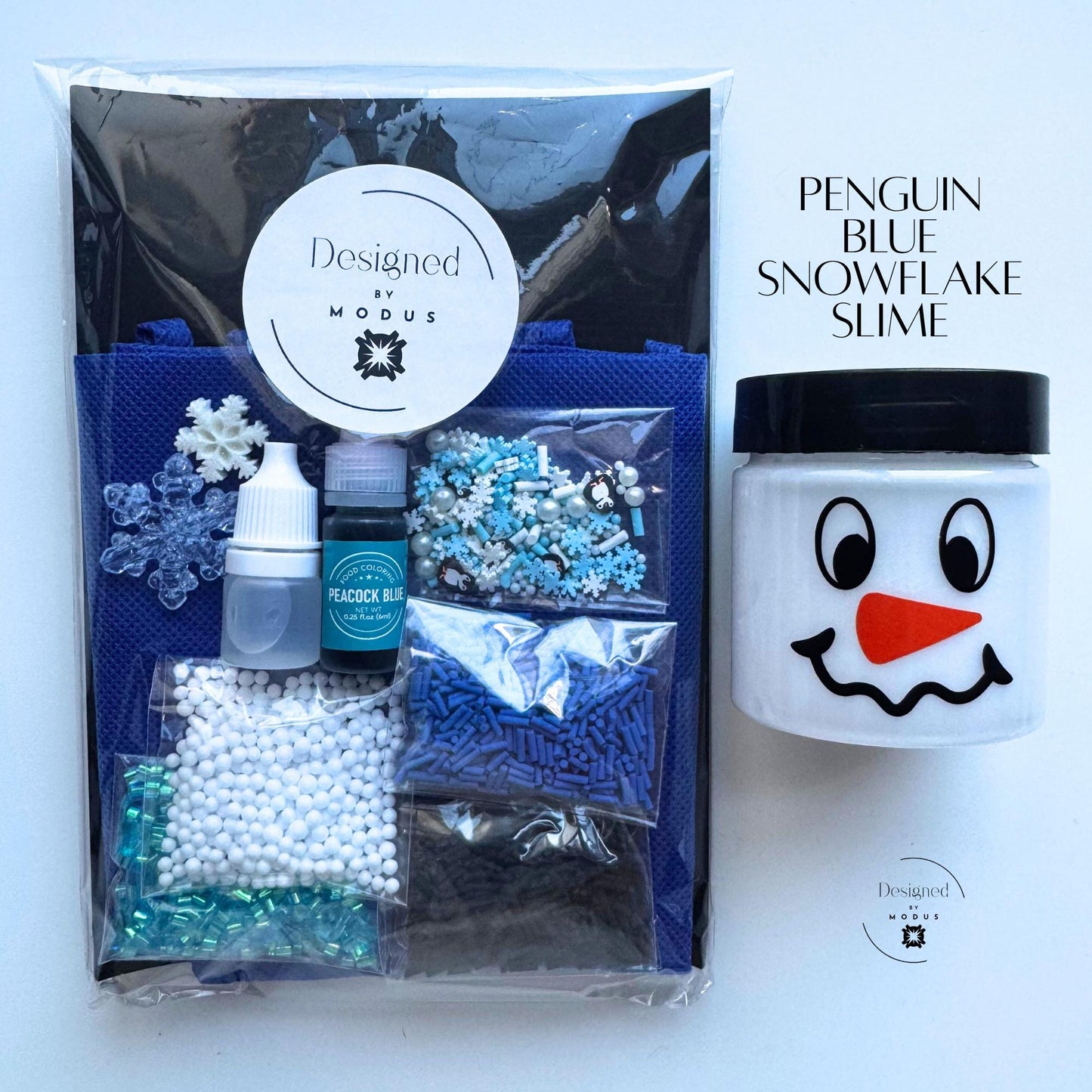 Packaged Penguin Blue Snowflake Slime Kit with Designed by Modus label, navy apron, snowman jar, and blue winter-themed mix-ins labeled “Penguin Blue Snowflake Slime.”