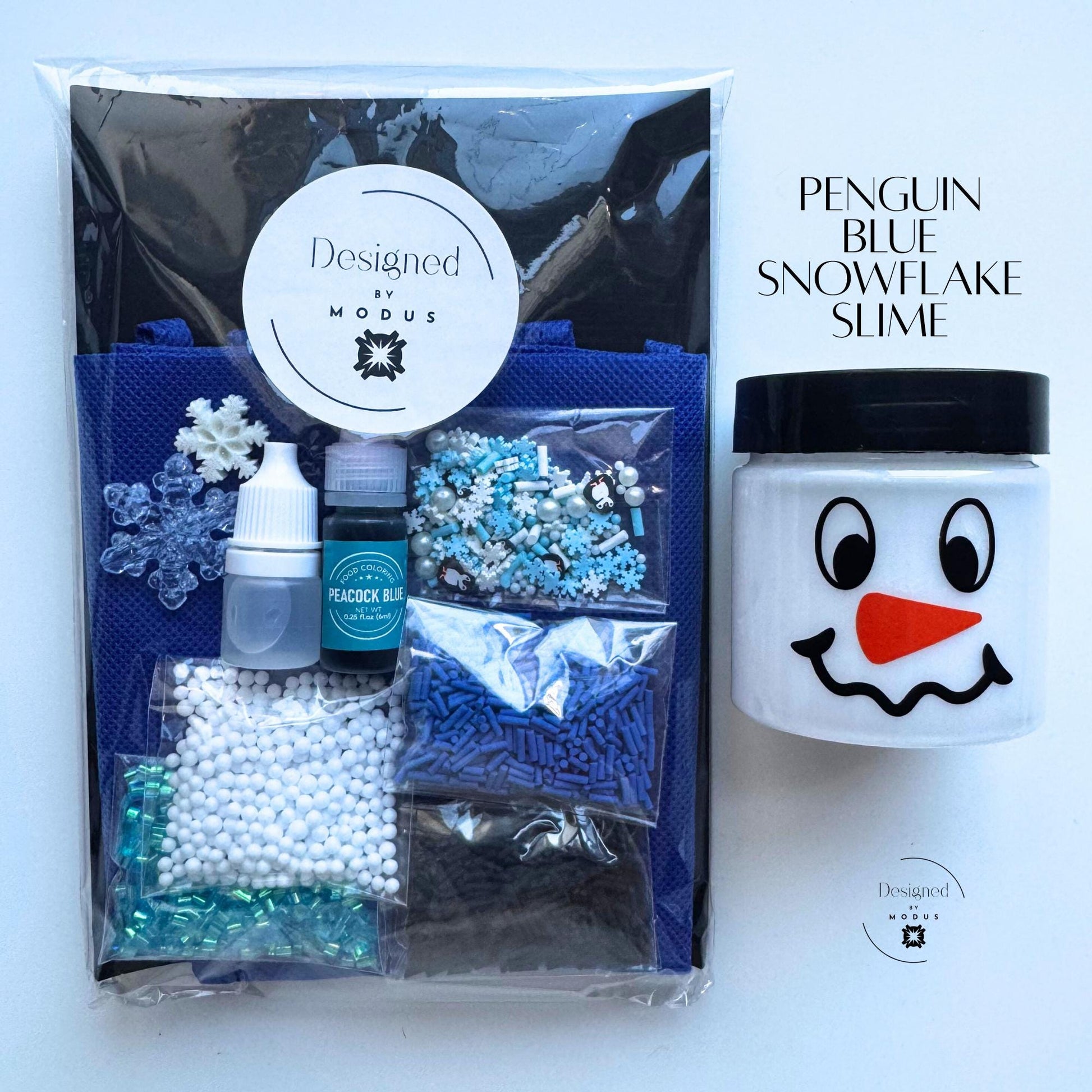 Packaged Penguin Blue Snowflake Slime Kit with Designed by Modus label, navy apron, snowman jar, and blue winter-themed mix-ins labeled “Penguin Blue Snowflake Slime.”