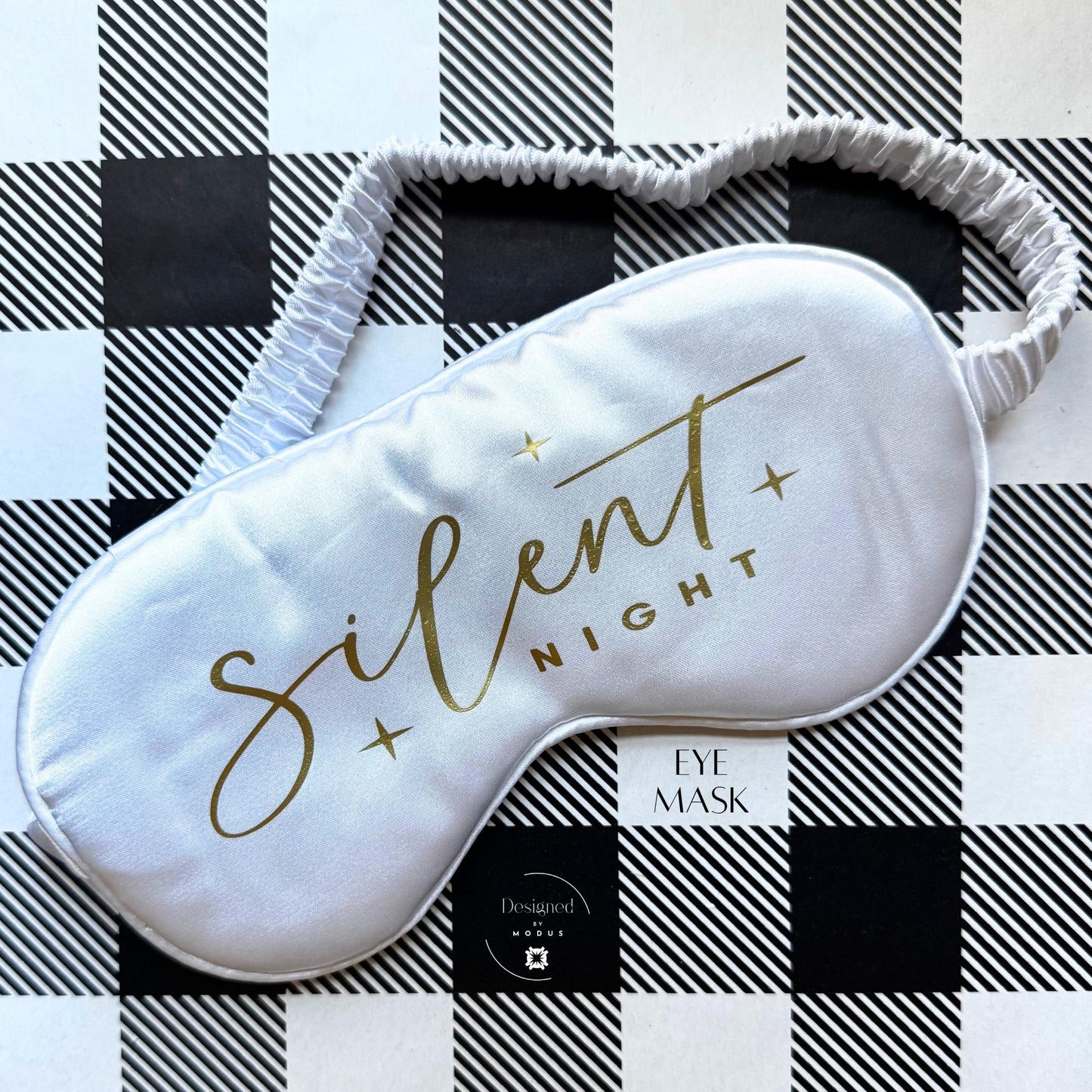 White satin eye mask with “Silent Night” printed in gold script on black-and-white plaid background.