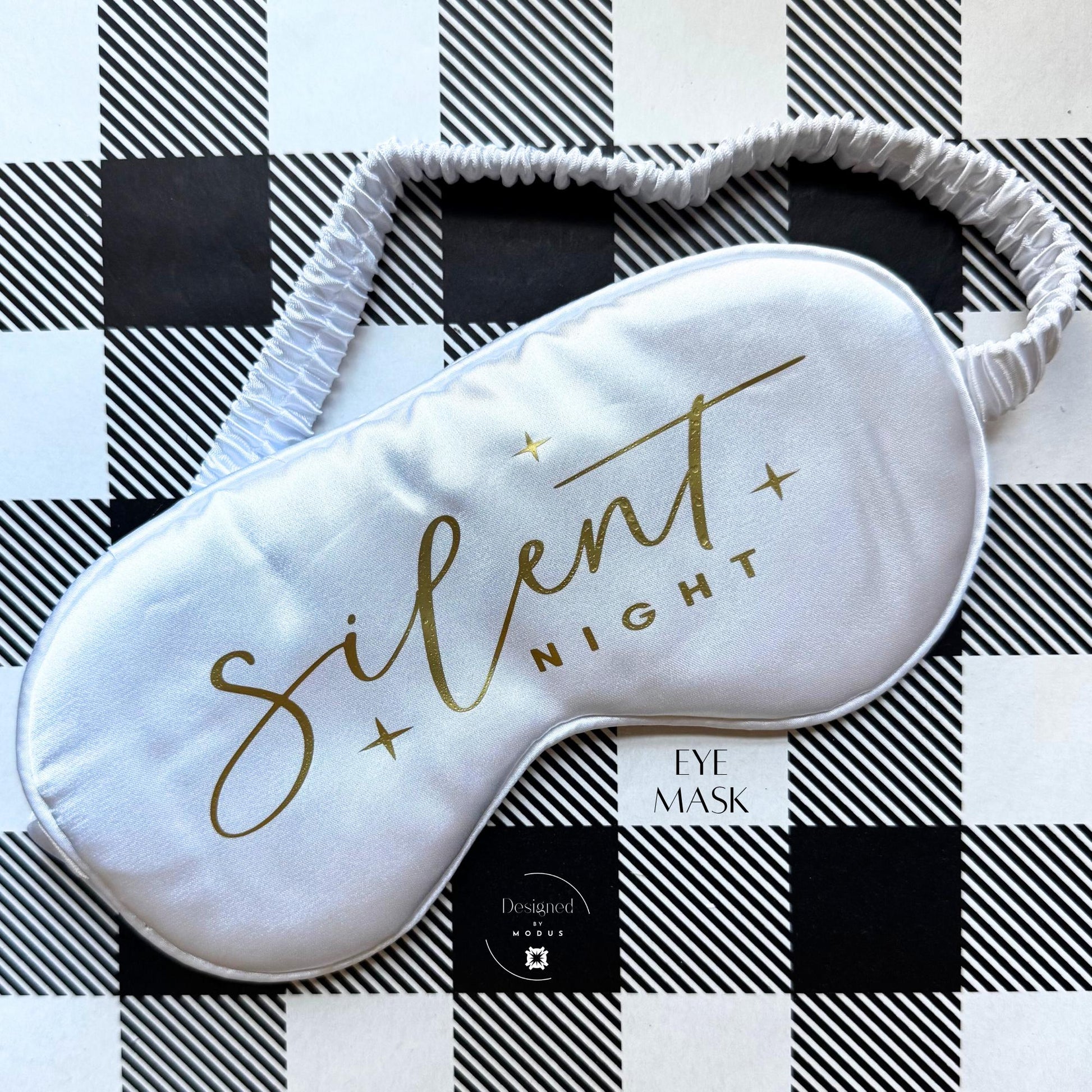 White satin eye mask with “Silent Night” printed in gold script on black-and-white plaid background.