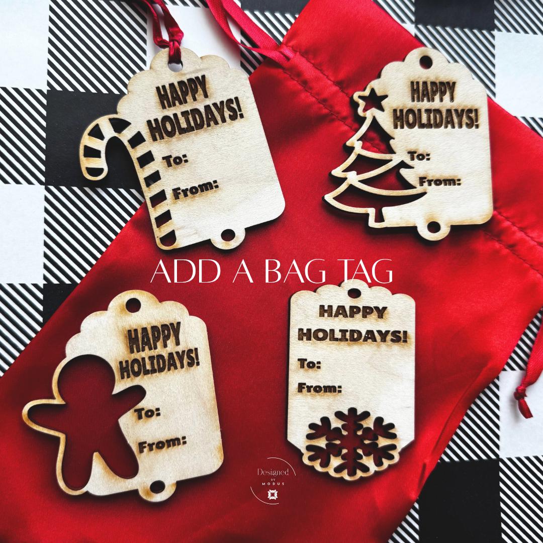 Four engraved wooden holiday tags — candy cane, tree, gingerbread, and snowflake shapes — displayed on red bag with caption “Add a Bag Tag.”