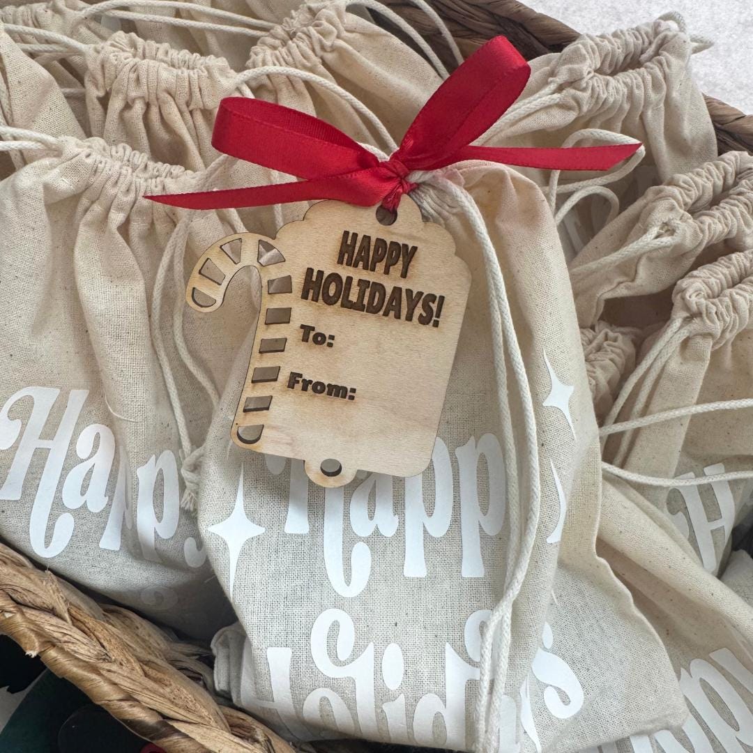 Candy cane-shaped wooden tag with engraved “Happy Holidays” message, tied to beige “Happy Holidays” bag with red ribbon, styled in festive basket.