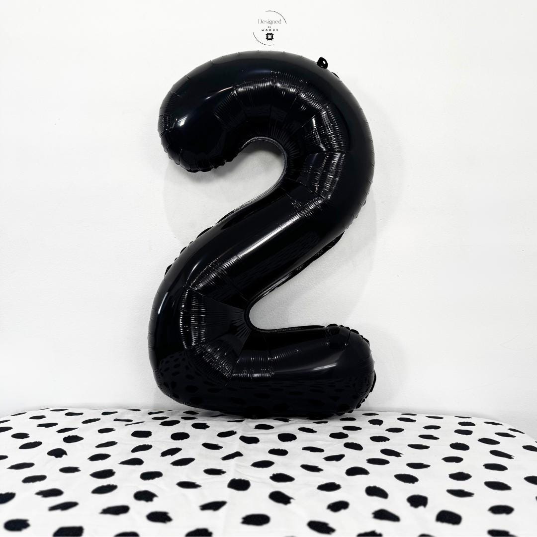 Shiny black number 2 foil balloon standing against white wall with black dotted surface.