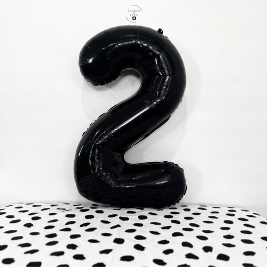 Shiny black number 2 foil balloon standing against white wall with black dotted surface.