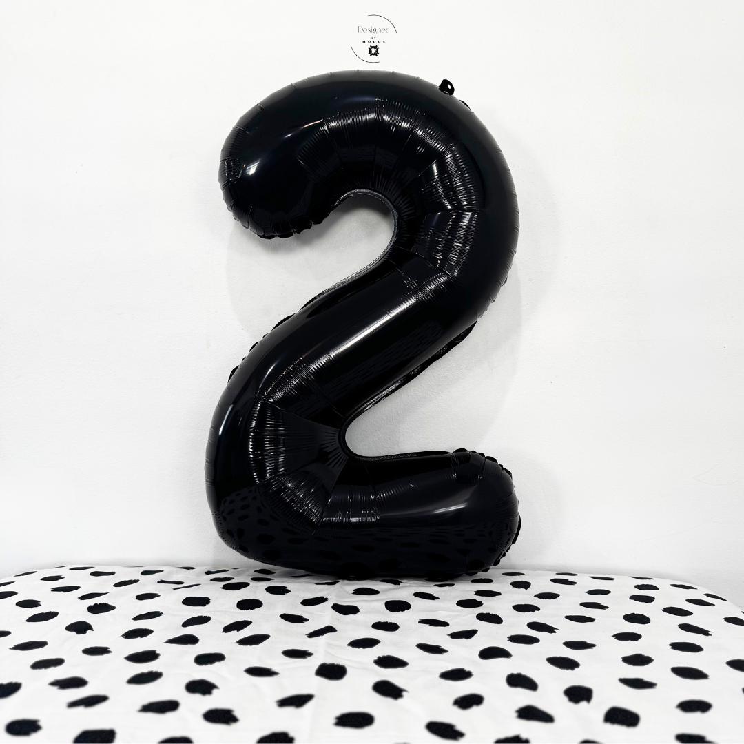 Shiny black number 2 foil balloon standing against white wall with black dotted surface.