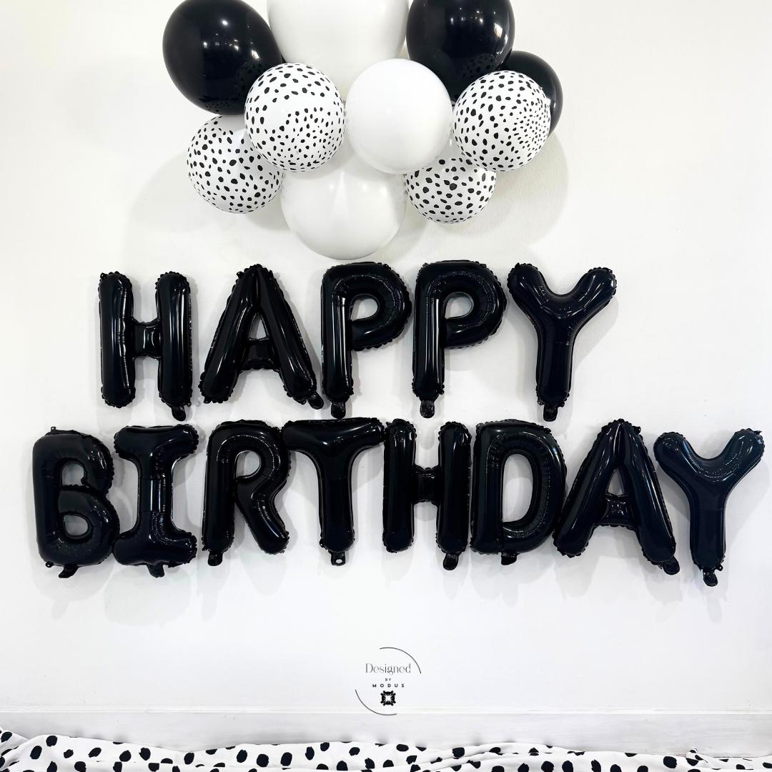 Black foil “HAPPY BIRTHDAY” balloon letters on white wall with black-and-white balloon cluster above.