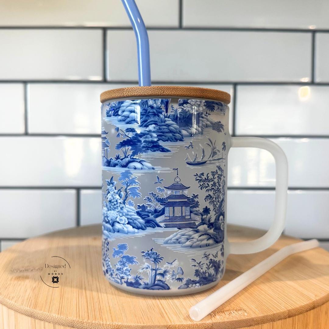 Frosted glass mug with blue and white chinoiserie design, bamboo lid, and straw on wood surface.