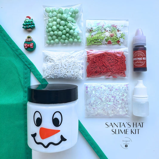 Santa’s Hat Slime Kit flat lay with green apron, snowman jar, and red, green, and white mix-ins labeled “Santa’s Hat Slime Kit.”
