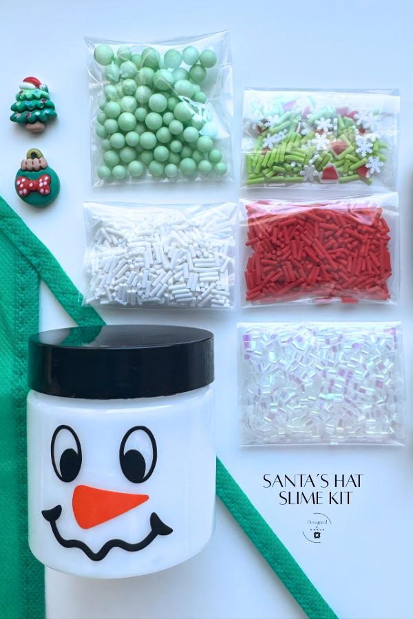 Santa’s Hat Slime Kit flat lay with green apron, snowman jar, and red, green, and white mix-ins labeled “Santa’s Hat Slime Kit.”