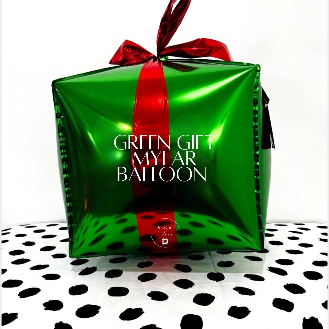 “Green metallic gift box mylar balloon with red ribbon on polka dot tablecloth for holiday display.”