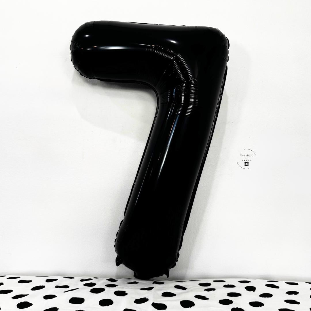 Shiny black number 7 foil balloon propped against minimalist white wall and dotted cloth.