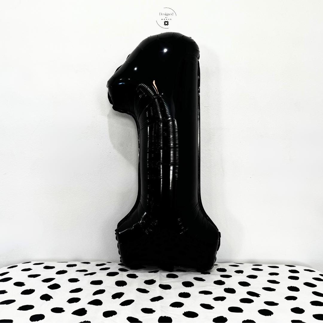 Large black number 1 foil balloon against white wall with black-and-white spotted backdrop.