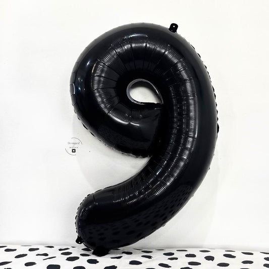 Large shiny black number 9 foil balloon against white wall and black-spotted fabric.