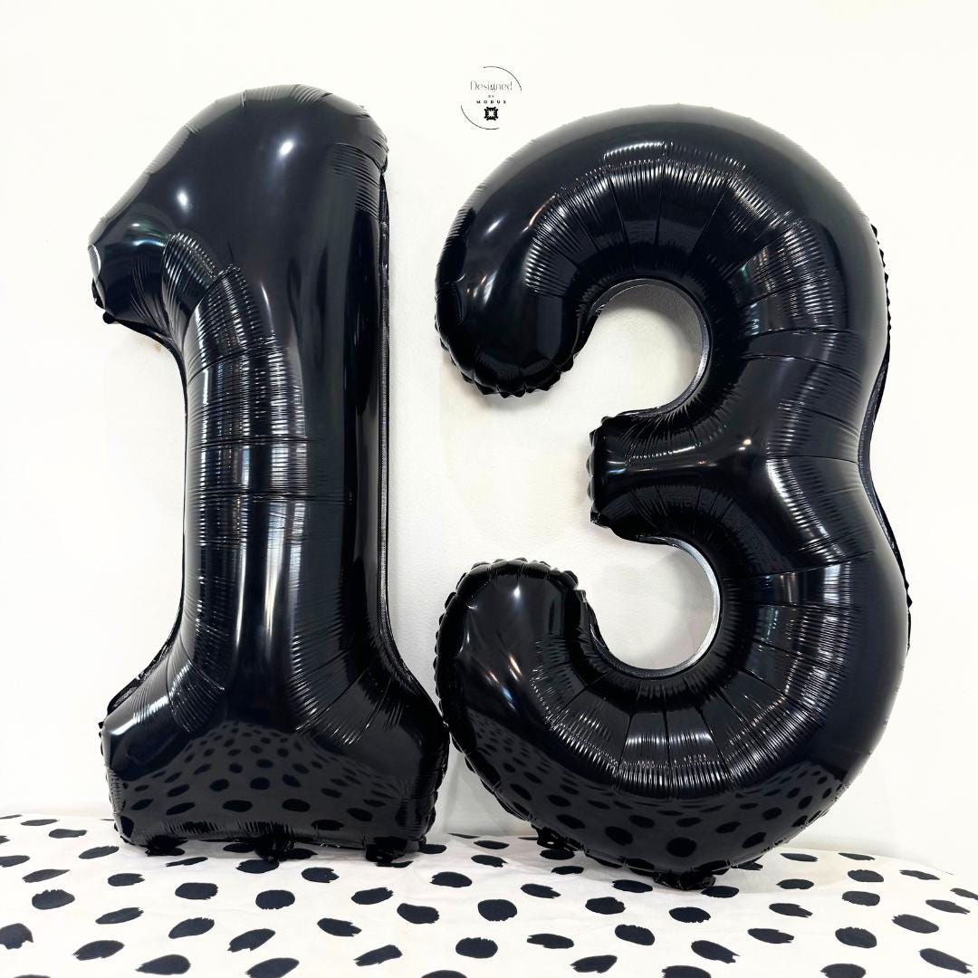 Glossy black number 13 foil balloons standing against white wall with black-dot fabric surface.