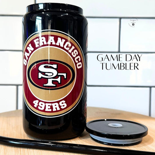 Black can-shaped tumbler with 49er red and gold football-inspired design and white straw on wood surface.