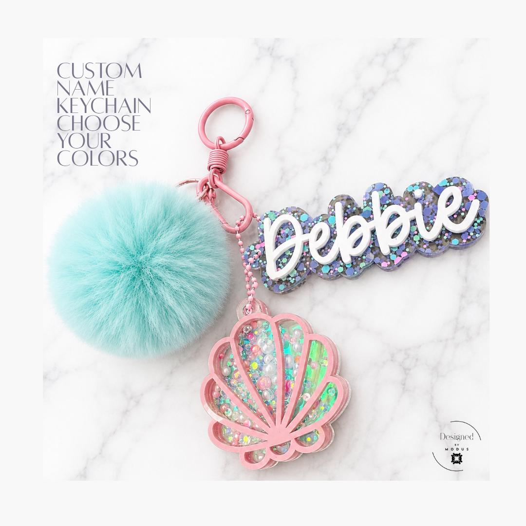 May include: A custom name keychain featuring the name Debbie in white script on a purple glitter background, a light blue fluffy pom-pom, and a pink shell-shaped charm filled with iridescent sequins, all attached to a pink metal ring.