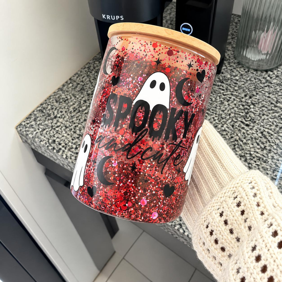 Cute ghost snow globe tumbler for Halloween featuring a clear cup filled with red, black and silver sparkling glitter. The front has a playful white ghost and spooky Halloween inspired graphic. Festive Halloween tumbler for enjoying seasonal drinks, decorating a Halloween party table or giving as a spooky season gift.